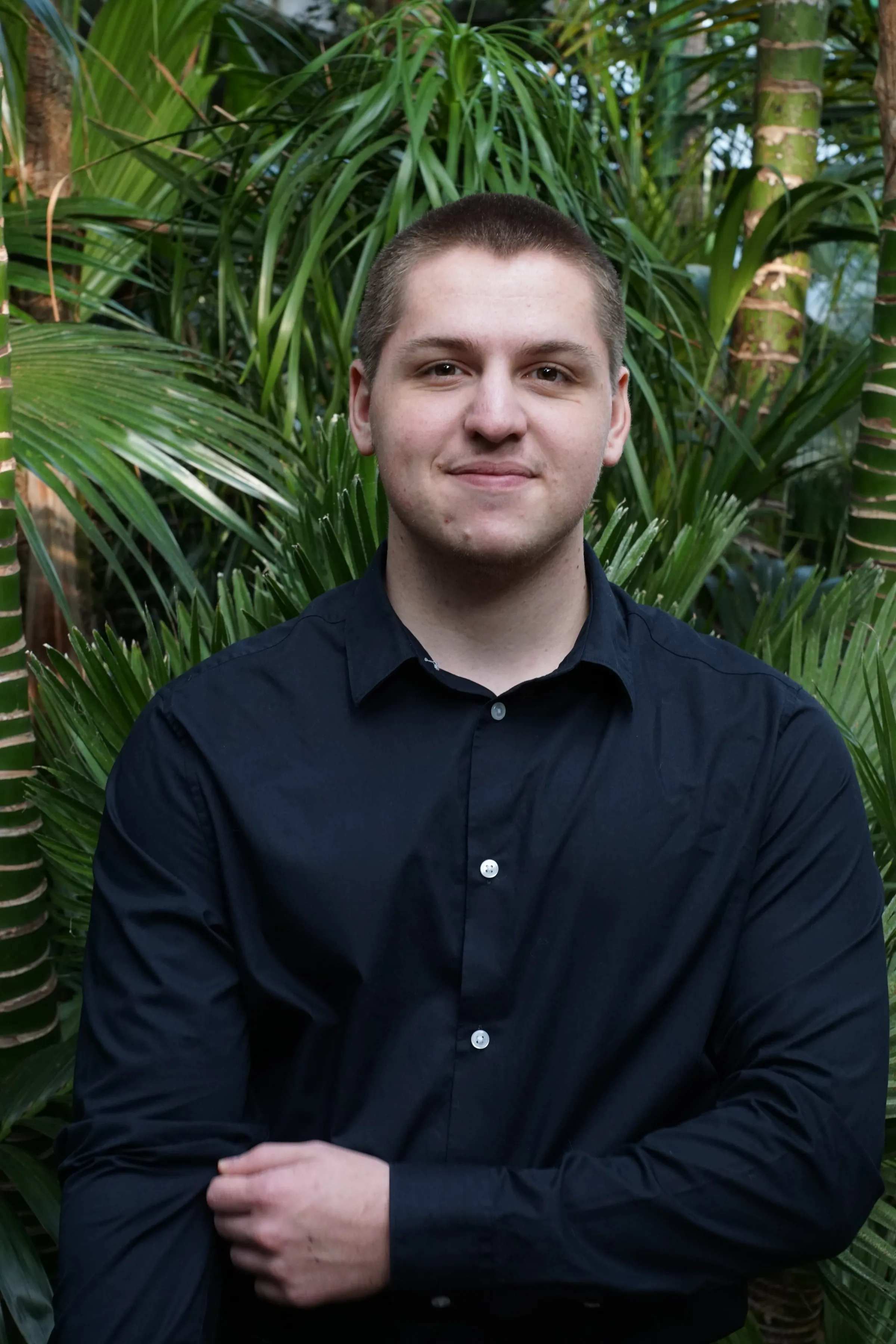 Kilian steht lächelnd mit verschränkten Händen in einem schwarzen Hemd vor grünen Pflanzen.