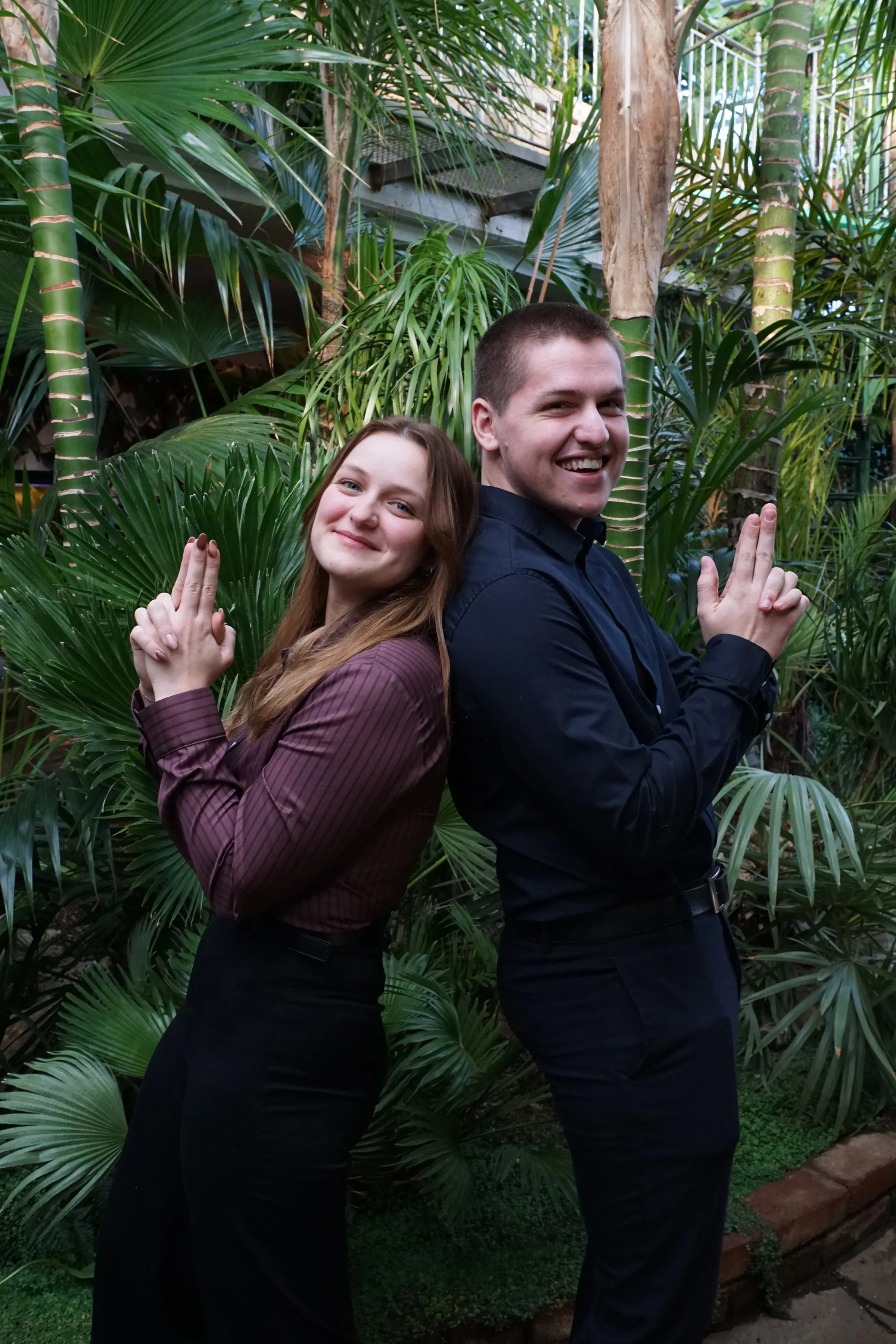 Kilian und Timea stehen Rücken an Rücken vor tropischen Pflanzen und posieren wie zwei Geheimagenten auf einer Mission.