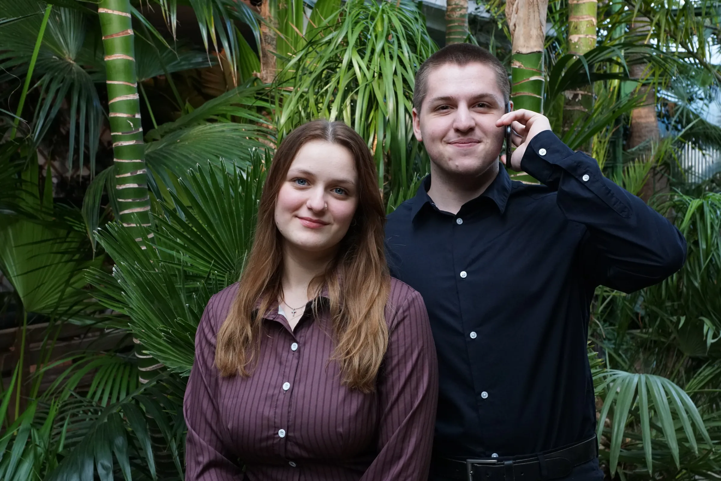 Kilian und Timea stehen nebeneinander vor einer Kulisse aus tropischen Pflanzen. Kilian hält ein Handy ans Ohr, während Timea freundlich in die Kamera lächelt.